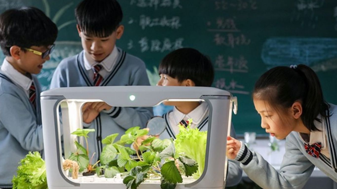 Four children in school uniforms gathered around a hydroponic system, observing leafy greens and strawberry plants under a grow light.